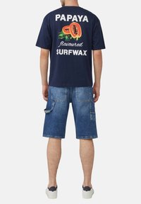 Person, der ein marineblaues T-Shirt mit Papaya-Grafik und Text, denimfarbene Cargo-Shorts und weiße Sneaker trägt, steht mit dem Rücken zur Kamera vor einem weißen Hintergrund.