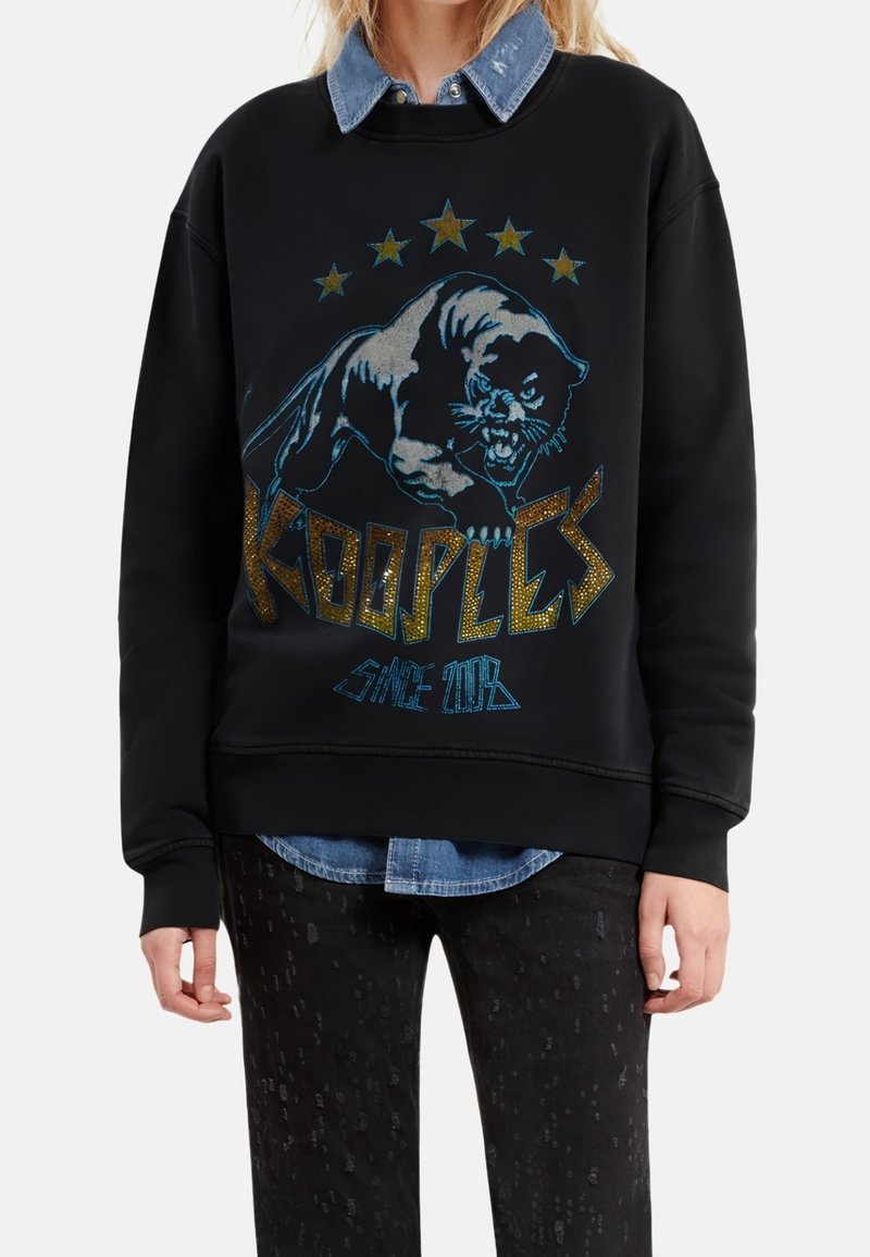 Person wearing black sweatshirt with roaring panther graphic, gold stars, and "Koooples Since 2008" text, over denim shirt and black pants.