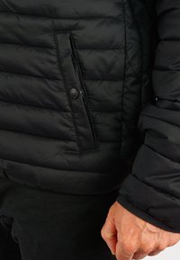 Črna puhovka s prešito zasnovo, ki ima gumbasto stransko žep in elastične man�šete. Tkanina ima gladek, sijajen videz.