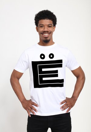 Hvid bomulds t-shirt med et stort sort grafisk design af bogstavet "E" og to cirkulære accenter, båret af en person med krøllet hår.