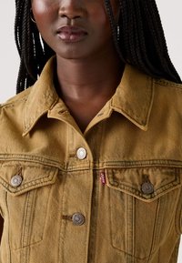 Dzeltenbrūna denima jaka ar divām krūšu kabatām, sudraba detaļām un Levi's etiķeti, ar klasisku apkakli un izturīgu tekstūru.