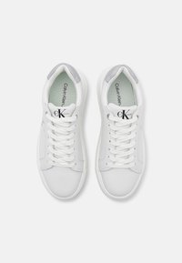 Bílé tenisky s hladkým koženým svrškem, plochými tkaničkami a světle šedými akcenty na patě. Obsahují texturovanou stélku a detaily s logem.