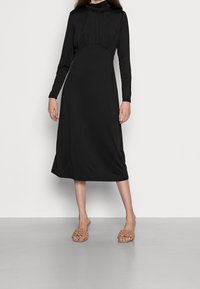Svart långärmad klänning med hög krage, rynkad liv, och rak silhuett. Bärs med ljusbruna texturerade sandaler.