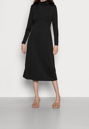 Vestido negro de manga larga con cuello alto, cuerpo fruncido y silueta recta. Combinado con sandalias de deslizar texturizadas en color marrón claro.