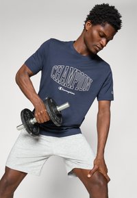 Marineblaue Champion-T-Shirt mit kurzen Ärmeln, das mit auffälligem weißen "CHAMPION"-Text versehen ist, kombiniert mit hellgrauen Sportshorts und einem schwarzen Hanteldumbbell.