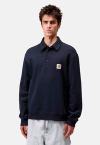 Suéter azul marino con cuello tipo polo, cuatro botones y un parche en el pecho. La tela parece suave, con un ajuste relajado y mangas largas.