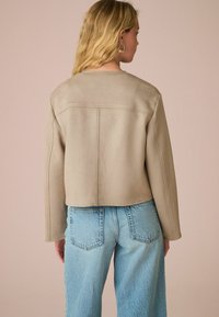 Veste cropped en faux daim beige avec des manches longues et un ourlet droit, associée à un jean large bleu clair, vue de dos.
