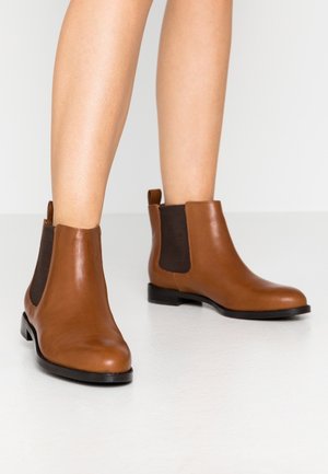 Bottes basses en cuir marron lisse avec panneaux latéraux élastiques foncés portées sur des jambes nues sur un fond clair uni.