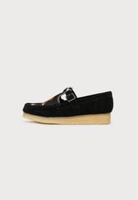 Fekete velúr slip-on cipő, lekerekített orral, díszítő csattal és mintás szőrme kiegészítővel a cipő elején. Gumitalp.