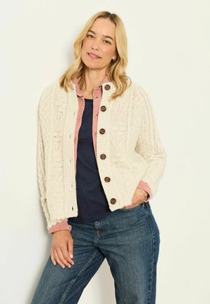 Woman with blond hair wearing a cream knit cardigan with brown buttons over a pink shirt and dark top, with blue jeans, standing against a white wall.