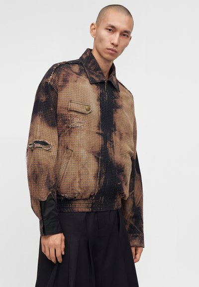 Άνδρας που φορά καφέ και μαύρο καρό μπουφάν με φθαρμένες λεπτομέρειες και μαύρο παντελόνι, στέκεται μπροστά σε απλό λευκό φόντο.