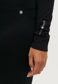 Zwarte lange mouwen onderlaag met een nauwsluitende pasvorm, zachte textuur en een logo op de mouw gedrukt. Naaddetail zichtbaar bij de pols.