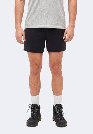 Homme portant un short noir, des chaussettes blanches mi-hautes, des chaussures de randonnée noires et un t-shirt gris, debout devant un fond clair uni.