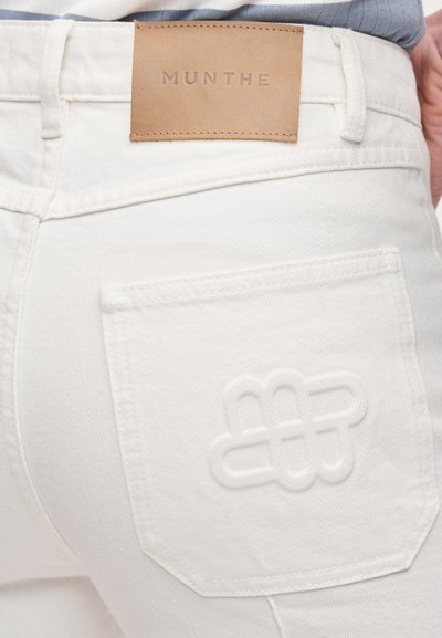 Λευκά τζιν κατασκευασμένα από υφασμάτινο βαμβάκι με υφή, με δερμάτινη ετικέτα που φέρει τη λέξη "MUNTHE" και ανάγλυφο μοτίβο στην πίσω τσέπη.