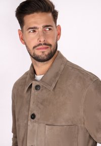 Jachetă din suede de culoare maro deschis, cu guler, închidere cu nasturi și buzunar frontal. Textură moale și design minimalist, cu o croială clasică.
