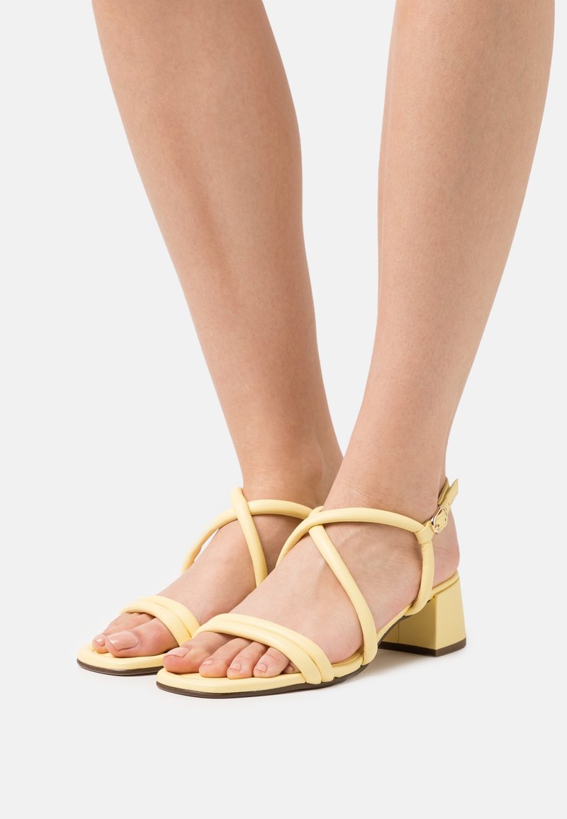 Jambes portant des sandales à brides jaune pâle avec des talons blocs bas sur un fond clair.