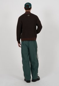 Brun strikkegenser med høy hals, kombinert med løse grønne bukser med hvite sidestriper. Sorte kraftige sko fullfører antrekket.