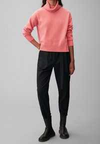 Persoon in een roze coltrui, zwarte hoge taille tapered broek met een zijstrik en zwarte enkellaarsjes, staande tegen een grijze achtergrond.