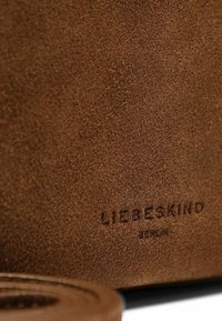 Il materiale scamosciato marrone con una texture morbida presenta il logo impresso "LIEBESKIND BERLIN". In primo piano è parzialmente visibile una tracolla in pelle arrotolata.