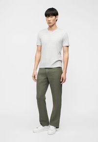 Ung mann stående, iført lysegrå kortermet V-hals skjorte, olivengrønne bukser med rette ben og hvite joggesko mot en ensfarget bakgrunn.