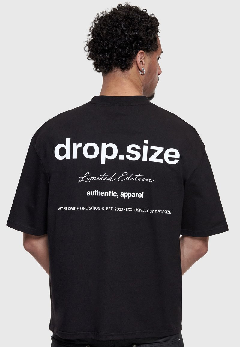 Homme aux cheveux bouclés vu de dos portant un t-shirt noir avec un texte blanc « drop.size Limited Edition authentic, apparel » dans le dos.