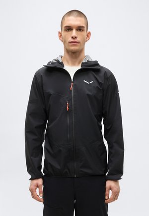 Jeune homme aux cheveux courts portant une veste légère noire avec capuche et une poche zippée sur la poitrine, debout devant un fond blanc uni.