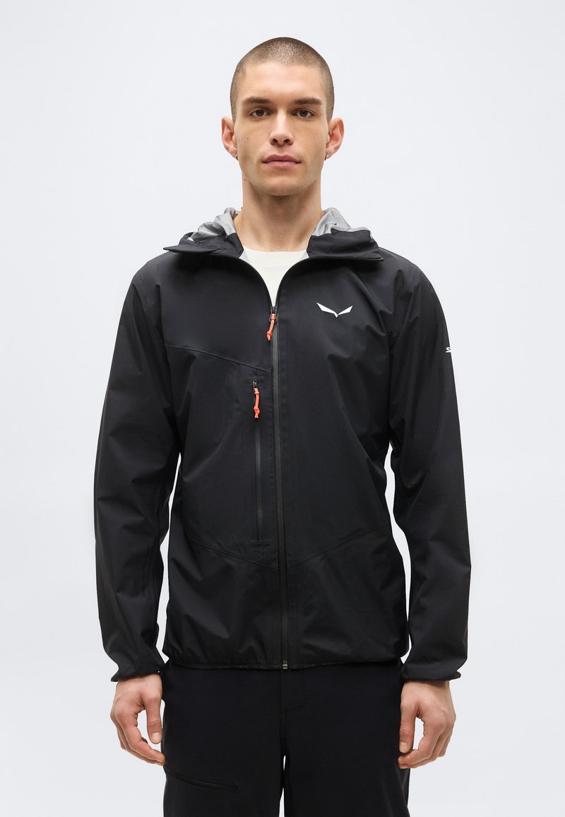 Jeune homme aux cheveux courts portant une veste légère noire avec capuche et une poche zippée sur la poitrine, debout devant un fond blanc uni.