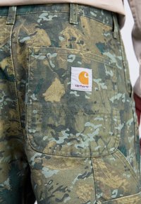 Pantaloni corti con motivo mimetico in tonalità terrose, con un'etichetta in tessuto che presenta un logo arancione. Dotati di una tasca posteriore con bordi arrotondati.