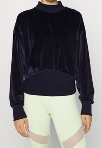 Sweatshirt en velours bleu marine avec un col montant, une coupe ample et des poignets côtelés. Caractérisé par une texture douce et un ourlet froncé. Associé à des leggings vert clair.