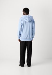 Hanorac albastru deschis cu glugă ajustabilă, croială lejeră și mansete ribbate, asortat cu pantaloni negri cu croială largă. Textură de țesătură netedă.