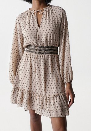 Kobieta w beżowej, długorękawowej sukience z małym czarnym wzorem, marszczonym pasem, wiązanym dekoltem i falbaniastym dołem na jednolitym tle.