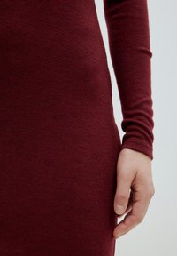 Bordeaux gebreid jurk met een aansluitend ontwerp, voorzien van een geribbelde textuur en lange mouwen, waarbij een deel van een hand nabij de zoom zichtbaar is.