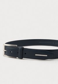 Tmavomodrý kožený opasok so striebornou sponou a kovovou štítkom značky Armani Exchange na konci, s textúrovaným povrchom, položený na svetlom pozadí.