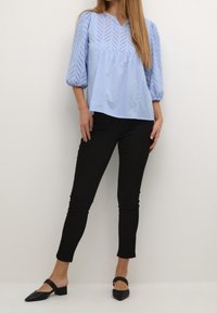 Ljusblå blus med ett chevron-perforerat mönster, samlade ärmar och en avslappnad passform, matchad med smala svarta byxor och svarta blockklackskor.