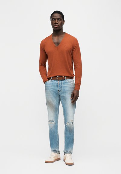 Homme portant un pull à manches longues couleur rouille avec col en V, jeans déchirés bleu clair, ceinture marron et baskets blanches, debout devant un fond uni.