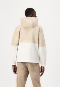 Beige and white hooded jacket with a matte finish, featuring a cinched hem and elastic cuffs. Back design includes a brand logo near the waist.