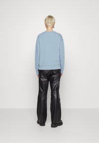 Una persona con capelli biondi corti si trova di fronte a una parete bianca, indossando una felpa azzurro chiaro, pantaloni di pelle nera e stivali neri.
