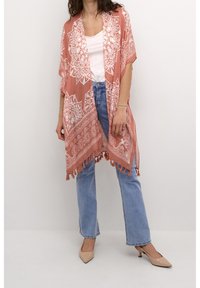 Lichtgewicht koraalkleurige kimono met witte mandalapatronen, franjes aan de zoom en een open voorkant. Gecombineerd met een basic witte top en blauwe jeans.