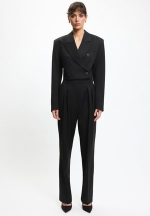 Kobieta stojąca w czarnym dwurzędowym skróconym blezerze i czarnych plisowanych spodniach z wysokim stanem oraz czarnych szpilkach z ostrym noskiem.