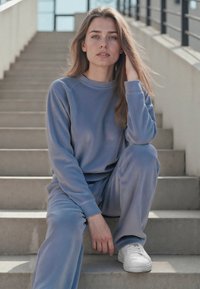 Completo tuta in tessuto morbido di colore blu chiaro, dal taglio rilassato, con maniche lunghe, scollatura a girocollo e pantaloni abbinati, indossato con scarpe da ginnastica bianche.