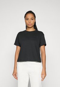 Moteris su pintomis plaukais, vilkinti paprastą juodą trumpą rankovę marškinėlį ir baltus kelnes, stovinti prieš paprastą šviesų foną.