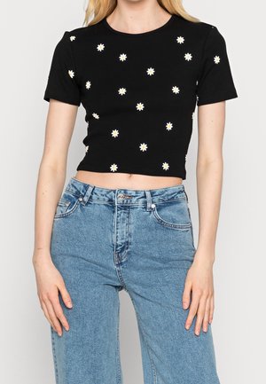 Mujer con una camiseta negra corta con pequeños patrones de margaritas blancas, combinada con jeans de tiro alto de color azul claro, de pie frente a un fondo liso.