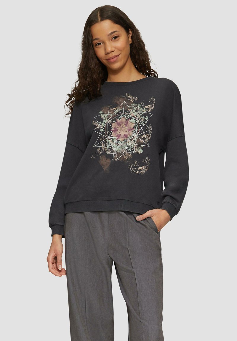 Czarna bluza z geometrycznym wzorem kwiatowym, z długimi rękawami i luźnym krojem, zestawiona z szarymi spodniami. Materiał wydaje się być miękki i textured.