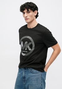 T-shirt de algodão preta com um logótipo circular cinza "MK" com riscas horizontais, emparelhada com jeans de ganga azuis.