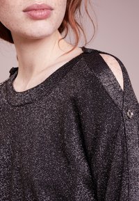 Maglione nero, in tessuto strutturato, con una finitura metallica lucida. Spalle con aperture distintive e dettaglio con bottoni sul collo.