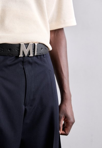 Black leather belt featuring a large silver 'M' buckle, worn over navy trousers. The outfit includes a textured cream top.