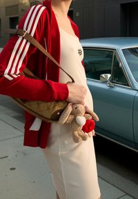 Personne portant une robe blanche et une veste rouge tenant un sac à main marron et un porte-clés lapin en peluche, debout à côté d'une voiture vintage bleue.