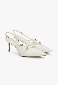 Escarpins slingback à bout pointu blanc cassé brillant, avec fines brides et détails de boucle dorée, talon stiletto de hauteur moyenne sur fond blanc.
