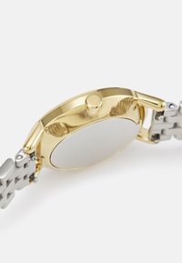 Montre avec boîtier doré et bracelet en acier inoxydable argent poli. Présente un dos rond et lisse ainsi qu'une petite couronne sur le côté.