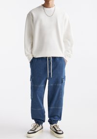 Vit sweatshirt med rund hals, kombinerad med blå denim cargo-byxor med elastiska mudd och dragsko i midjan. Svarta och gräddfärgade skor.